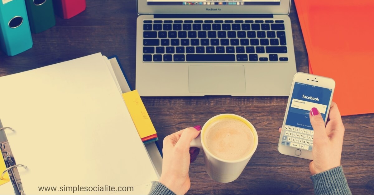 Woman holding coffee while on Facebook on her phone, in front of her laptop